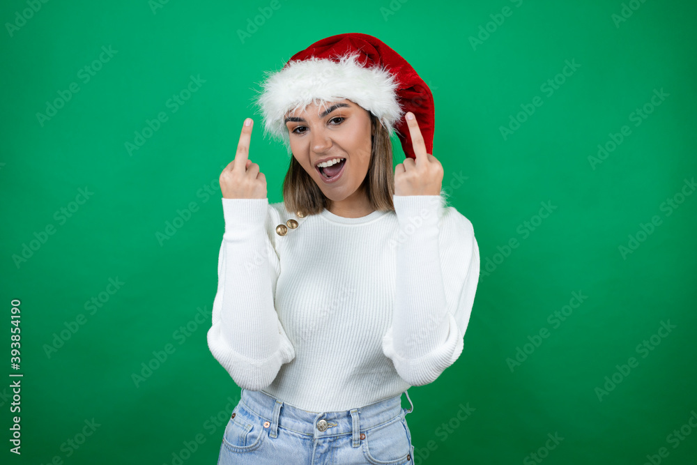 Young beautiful woman wearing a Santa hat and white sweater over green background showing middle finger doing fuck you bad expression, provocation and rude attitude. screaming excited Young beautiful woman wearing a Santa hat and white sweater over green background showing middle finger doing fuck you bad expression, provocation and rude attitude. screaming excited