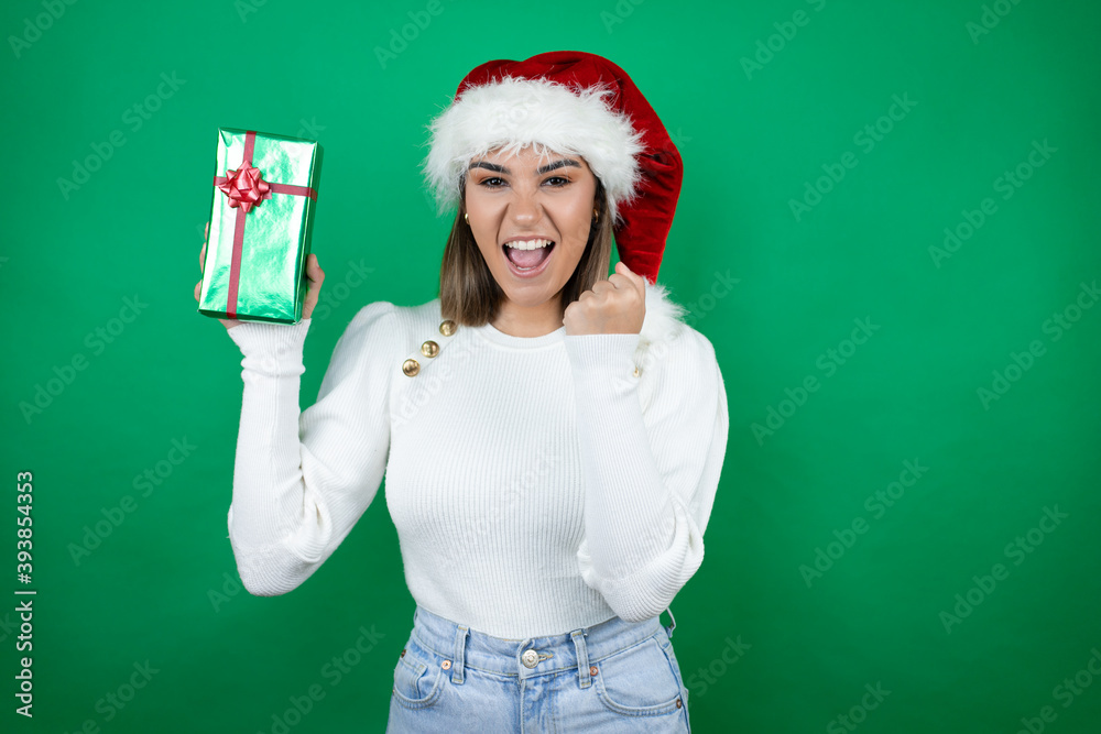 Young beautiful woman wearing a Santa hat holding a gift and white sweater over green background very happy and excited making winner gesture with raised arms, smiling and screaming for success. Young beautiful woman wearing a Santa hat holding a gift and white sweater over green background very happy and excited making winner gesture with raised arms, smiling and screaming for success.