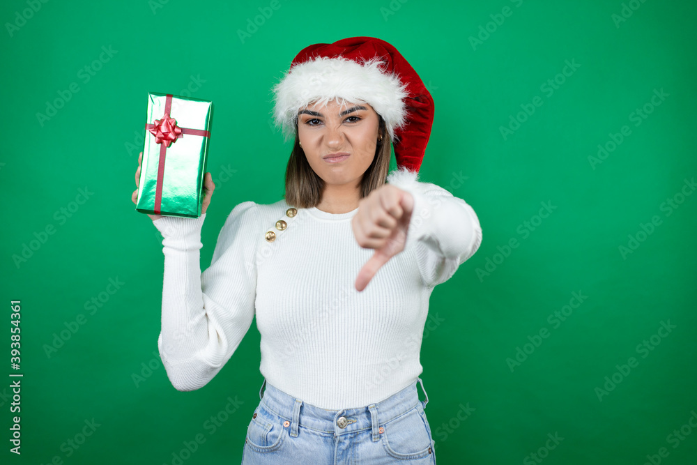 Young beautiful woman wearing a Santa hat holding a gift and white sweater over green background with angry face, negative sign showing dislike with thumb down Young beautiful woman wearing a Santa hat holding a gift and white sweater over green background with angry face, negative sign showing dislike with thumb down