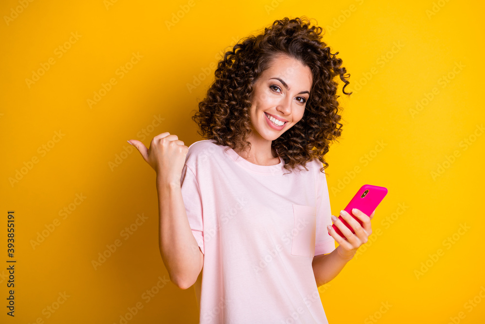 Photo portrait of curly curious girl keeping cellphone pointing at empty space isolated on vibrant yellow color background Photo portrait of curly curious girl keeping cellphone pointing at empty space isolated on vibrant yellow color background