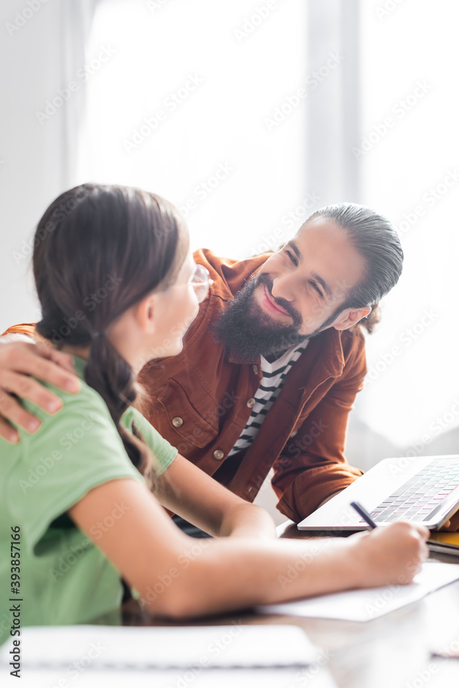 Happy hispanic father with laptop embracing daughter doing homework in kitchen on blurred foreground Happy hispanic father with laptop embracing daughter doing homework in kitchen on blurred foreground