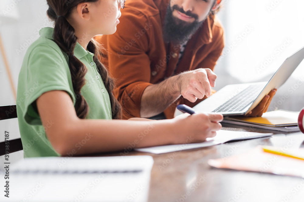 Cropped view of hispanic father pointing with finger at copybook near daughter doing homework on blurred foreground Cropped view of hispanic father pointing with finger at copybook near daughter doing homework on blurred foreground