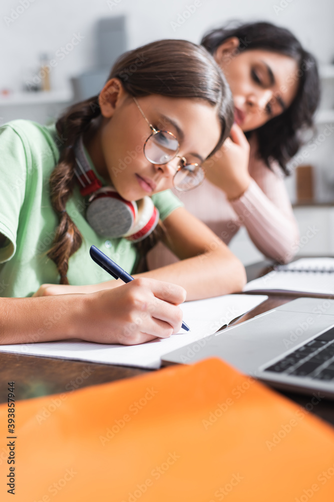 Hisapnic girl writing on notebook near laptop and mother on blurred background Hisapnic girl writing on notebook near laptop and mother on blurred background