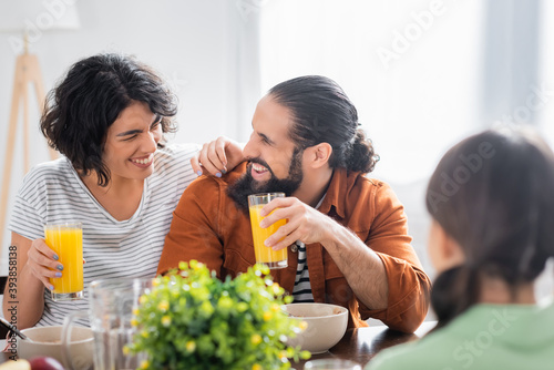 Cheerful hispanic parents with glasses of orange juice sitting near daughter on blurred foreground