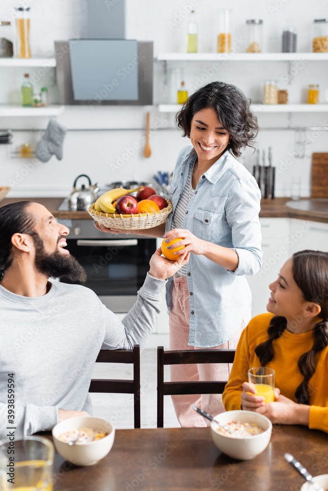 Smiling hispanic woman giving orange to husband near daughter and cereals on blurred foreground