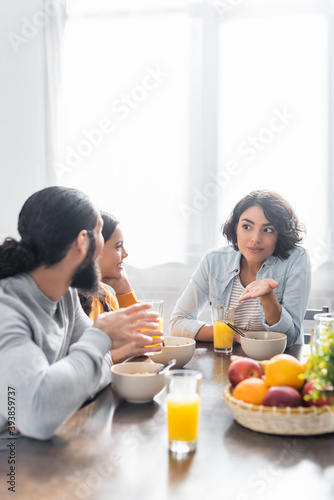 Hispanic woman pointing with hand near family and breakfast on blurred foreground in kitchen