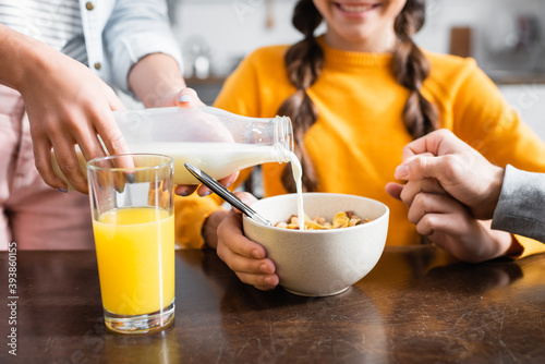 Cropped view of woman pouring milk in cereals near glass of orange juice and family on blurred foreground