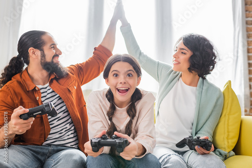 KYIV, UKRAINE - NOVEMBER 16, 2020: happy hispanic couple giving high five while playing video game with excited daughter