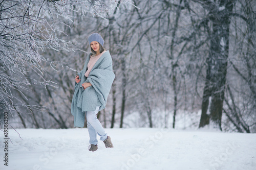 Beautiful woman rejoices in the first snow in winter. Snowy forest park. Walking with a blanket. Cold weather. Carelessness naivety. Light tones blue. Cozy time of the year. January December February