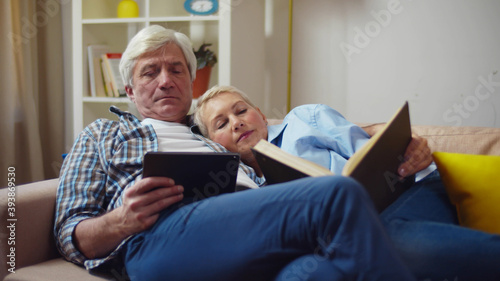 Senior couple reading book and using tablet on couch in living room