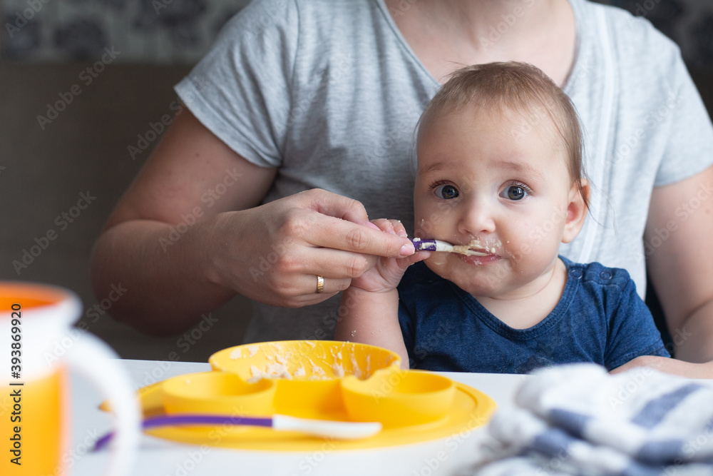 Cute infant trying to eat baby food using spoon and his cheerful mother assisting him. Cute infant trying to eat baby food using spoon and his cheerful mother assisting him.