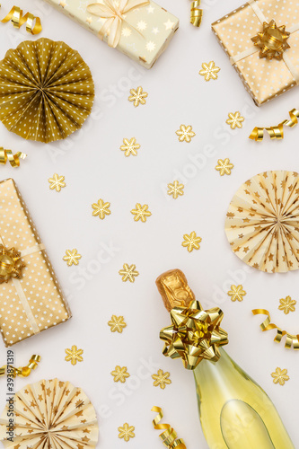 Festive white background with gold decoration , gift boxes with bottle of sparkling wine, shiny golden serpentine confetti and paper christmas tree decorations, glittering snowflakes, copy space