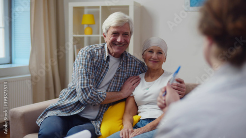 Female doctor visiting aged woman with cancer sitting on couch with husband photo