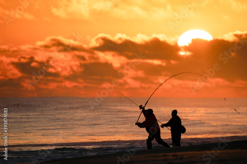 Silhouettes of surf casters fishing on the beach during a vibrant sunset. 
