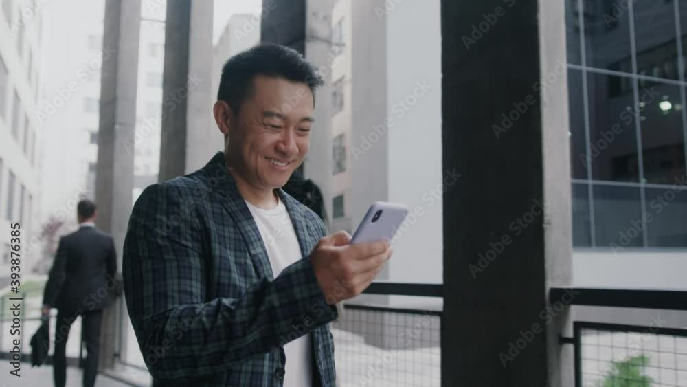 Happy asian young entrepreneur browsing mobile phone internet during office break walking along business district. Successful businessman going to work.