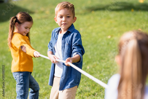 Boy holding rope while playing tug of war with friends on blurred foreground in park