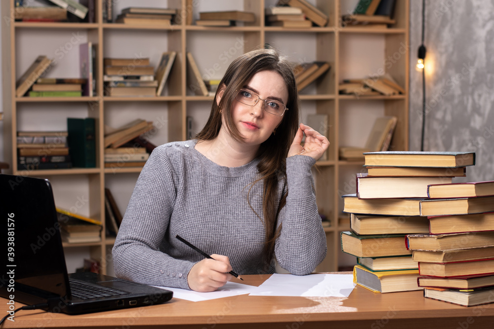 young student prepares for lessons. office worker writes in documents. a laptop stands on the table and a bookcase in the background. freelancer at work. look at the camera young student prepares for lessons. office worker writes in documents. a laptop stands on the table and a bookcase in the background. freelancer at work. look at the camera