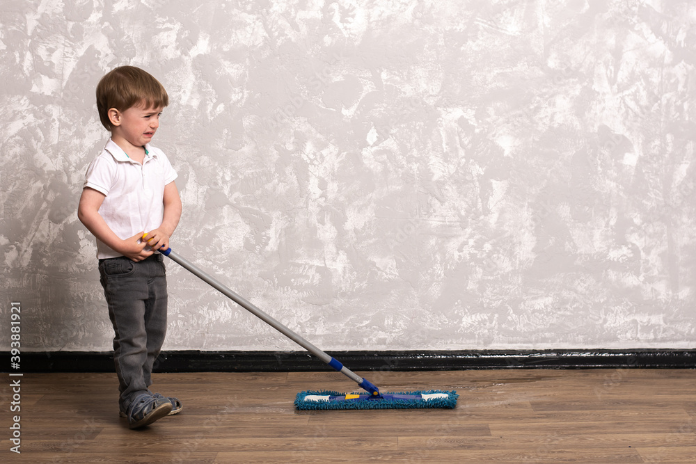 little boy washes the floor in the apartment with a mop. General cleaning of the house with the whole family. the child helps the parents. gray wall in the background. little boy washes the floor in the apartment with a mop. General cleaning of the house with the whole family. the child helps the parents. gray wall in the background.