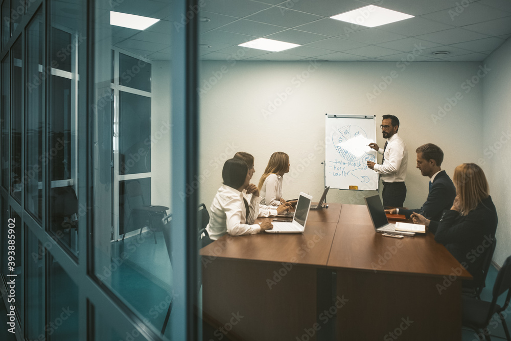 Business presentation concept. Speaker man stands near whiteboard having discussion with business people colleagues who are sitting at office table in office.