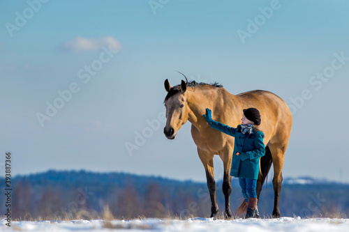 Beautiful girl standing near her Trakehner yellow horse outside in field. Child with a big mare standing together on snow in winter background