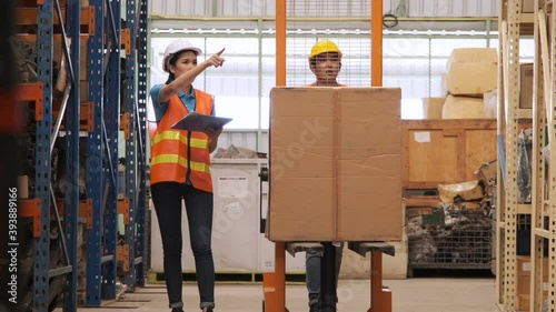 Asian woman manager hold clipboard for  checking list inventory with worker using hand stacker walking together in warehouse.