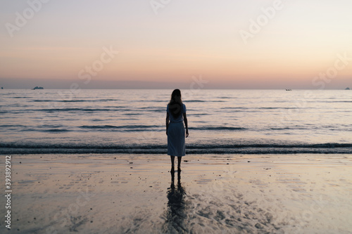 woman stares admires looking out to the sea from a beach at sunset with blue and orange night evening sky sarong long hair relaxing viewing 