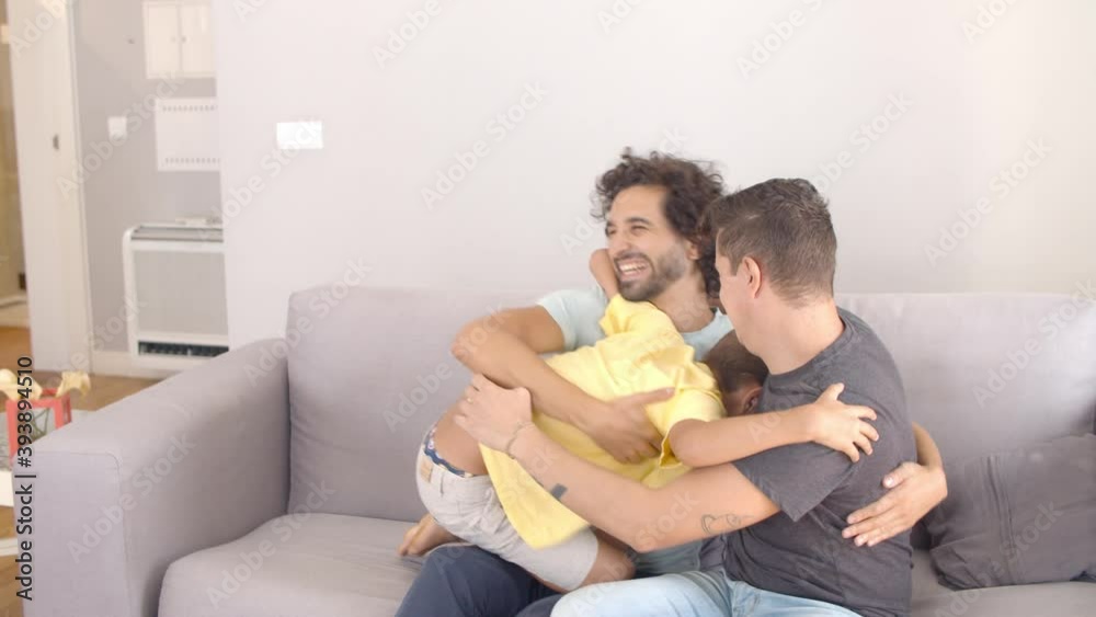 Cheerful boy jumping down on couch to his two dads and hugging them. Static shot. Parenthood and family concept Cheerful boy jumping down on couch to his two dads and hugging them. Static shot. Parenthood and family concept