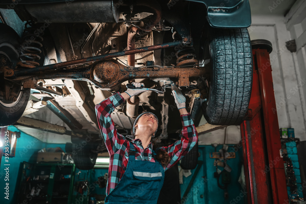 Obraz premium Portrait of a young female mechanic in uniform and gloves, repairing a car. Bottom view. The car is located on the lift