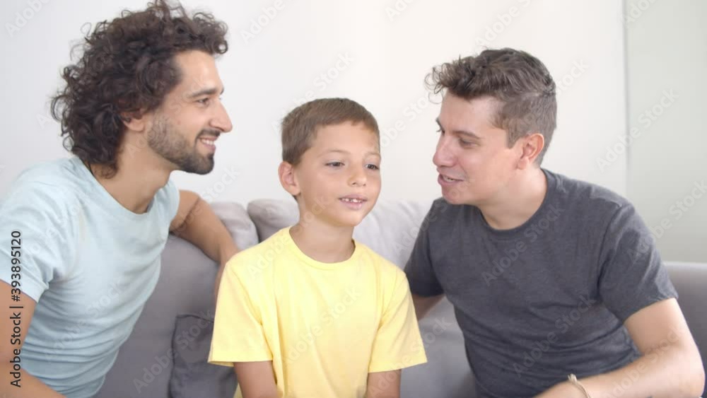 Two happy dads kissing son. Family sitting on couch, enjoying time together. Front view. Parenthood and love concept Two happy dads kissing son. Family sitting on couch, enjoying time together. Front view. Parenthood and love concept