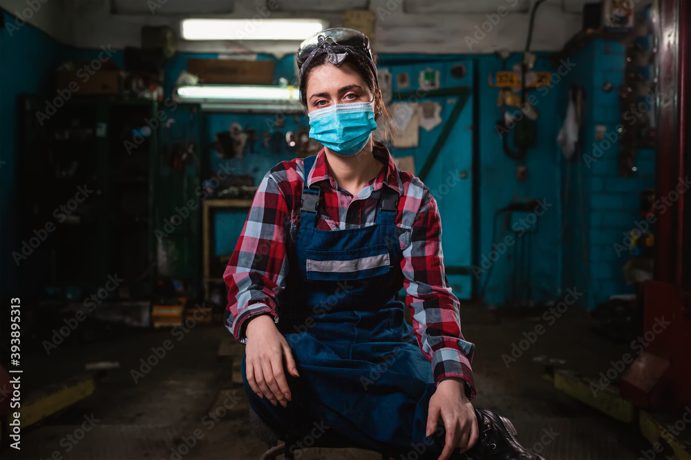 Portrait of a young female pretty mechanic in a work uniform is sitting on a chair, with a medical mask on her face. Indoor. Garage. Concept of virus protection in the workplace