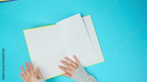 two female hands turn over the pages of a yellow notebook with blank white sheets on a blue background, top view