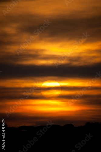 A setting sun behind layers of cloud and deep orange sky tones with a black horizon.Vertical Image