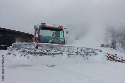 A snow plough sits waiting to continue clearing snow in a ski resort.In distance a ski slope ac be seen as snow falls