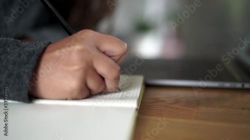 A person in the house working on her laptop and taking notes on a paper notebook.