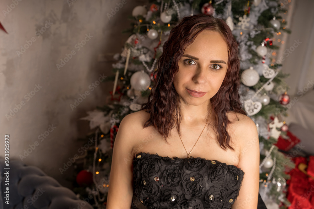 Portrait of charming brunette in black dress near Christmas tree. Young attractive woman posing at coniferous tree with decorative adornments. Concept of Christmas celebration at home.