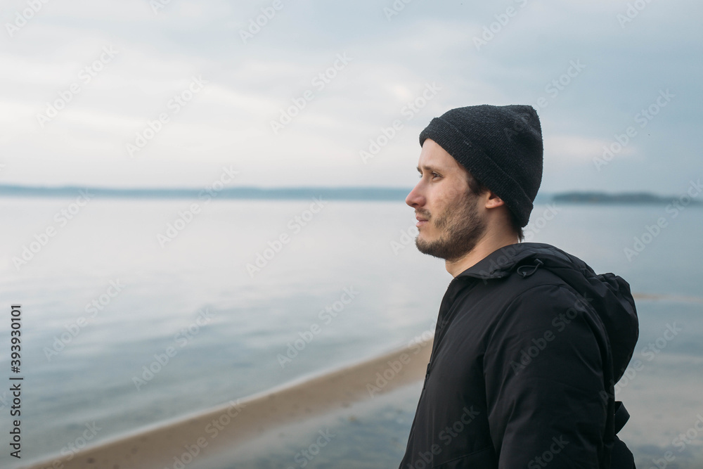 A portrait of a young guy in a black hat and black jacket looks thoughtfully into the distance against the background of the lake and the sky in profile.