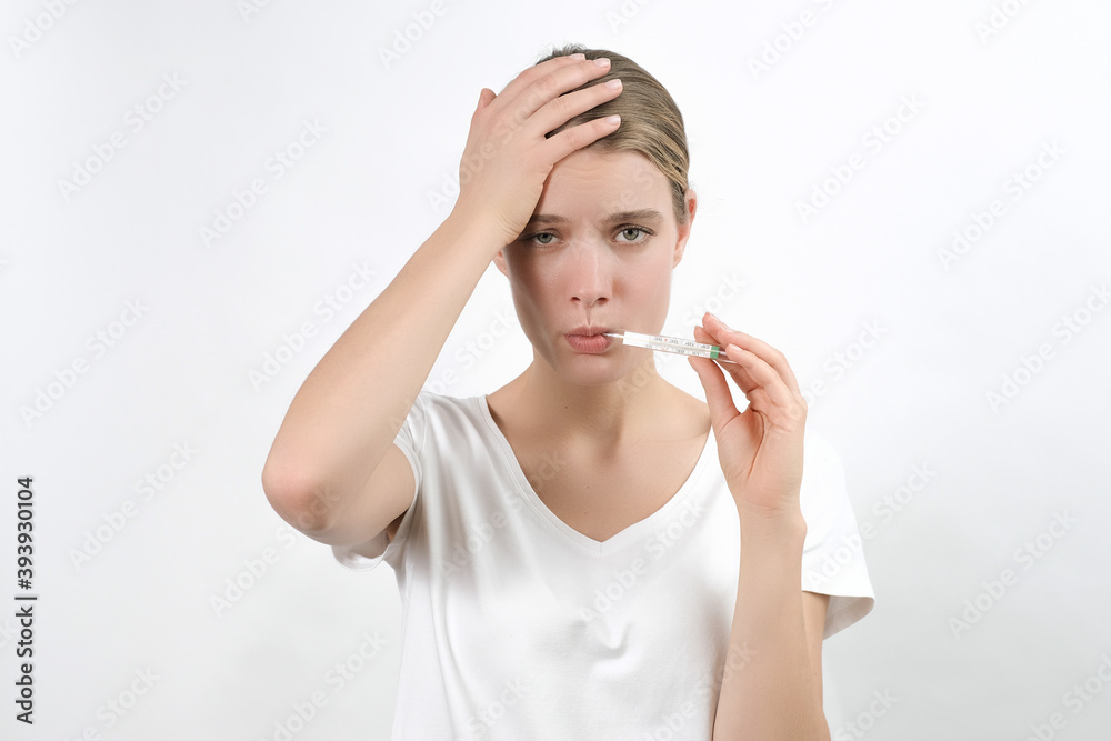 young caucasian blonde woman holding a thermometer in her mouth and keeping raised hand on her head and frowning sadly face, isolated on the white background. young caucasian blonde woman holding a thermometer in her mouth and keeping raised hand on her head and frowning sadly face, isolated on the white background.
