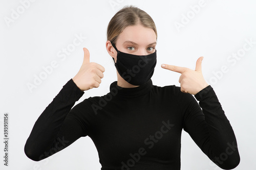 Young white woman wearing black face mask showing. thumb, ok sign and pointing with the index finger. Virus, covid-19 protection.