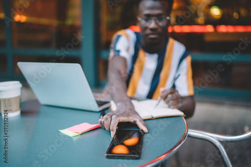 Selective focus on black male hand touching smartphone gadget for checking time during distance e learning in street cafe, blurred man using cellphone device for reading web news and texting