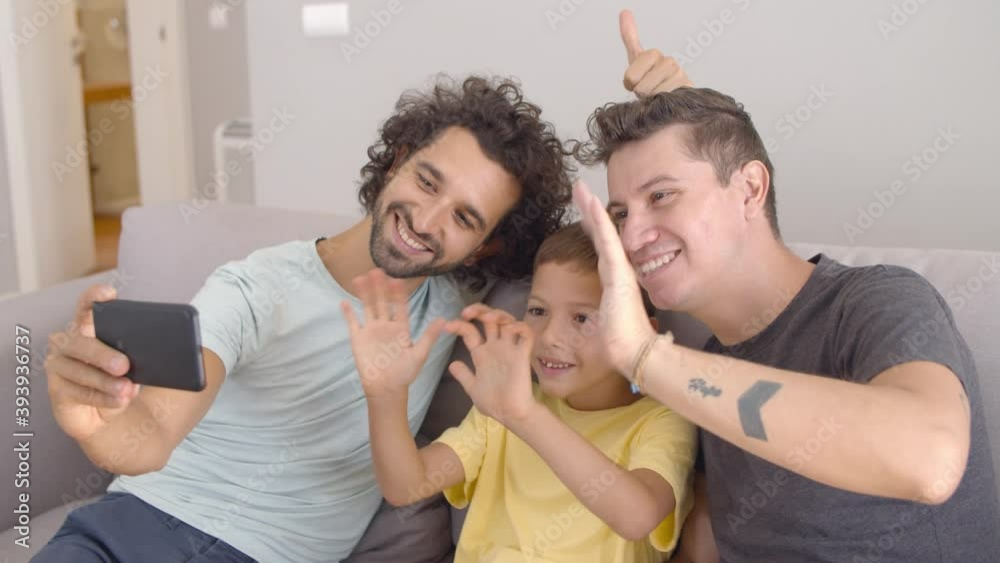 Two happy fathers and kid using cell for video call, waving hello at webcam, making like gesture and smiling at screen. Happy family and communication concept Two happy fathers and kid using cell for video call, waving hello at webcam, making like gesture and smiling at screen. Happy family and communication concept