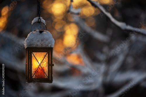 Candle lantern hanging from tree branch, bokeh winter background.