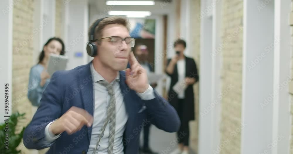 Joyful young businessman dancing in headphones as blurred colleagues recording video on gadgets at the background. Coworkers filming happy employee in office corridor. Cinema 4k ProRes HQ. Joyful young businessman dancing in headphones as blurred colleagues recording video on gadgets at the background. Coworkers filming happy employee in office corridor. Cinema 4k ProRes HQ.