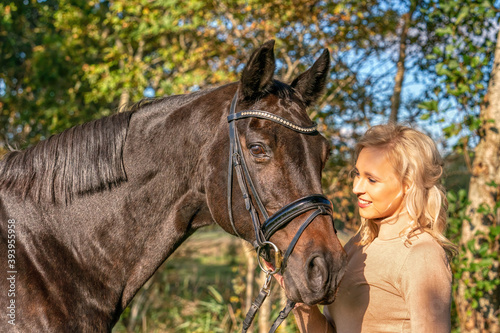 Side view, portrait. Female in casual clothes stands in the forest with her horse. In the autumn sun