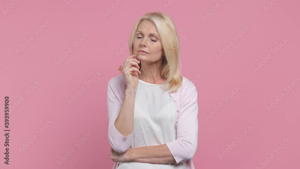 Thoughtful pensive elderly gray-haired blonde woman lady 40s 50s years old in white blank casual t-shirt prop up chin with hand think about something important isolated on pastel pink color background Thoughtful pensive elderly gray-haired blonde woman lady 40s 50s years old in white blank casual t-shirt prop up chin with hand think about something important isolated on pastel pink color background
