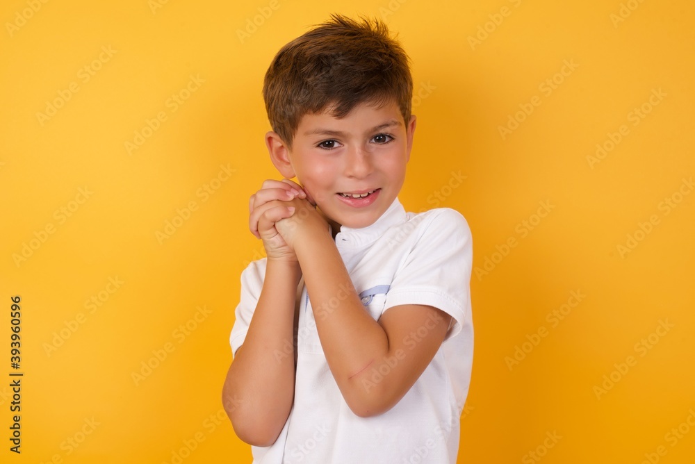 Dreamy little cute Caucasian boy kid wearing white t-shirt against yellow wall with pleasant expression, closes eyes, keeps hands crossed near face, thinks about something pleasant Dreamy little cute Caucasian boy kid wearing white t-shirt against yellow wall with pleasant expression, closes eyes, keeps hands crossed near face, thinks about something pleasant