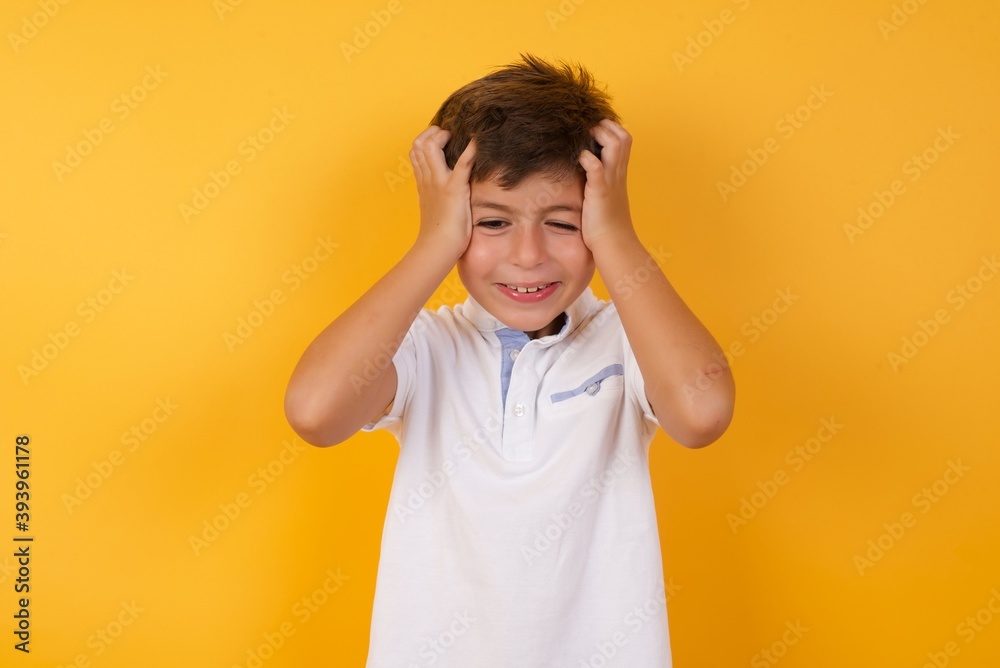 Shocked panic little cute Caucasian boy kid wearing white t-shirt against yellow wall holding hands on head and screaming in despair and frustration. Shocked panic little cute Caucasian boy kid wearing white t-shirt against yellow wall holding hands on head and screaming in despair and frustration.