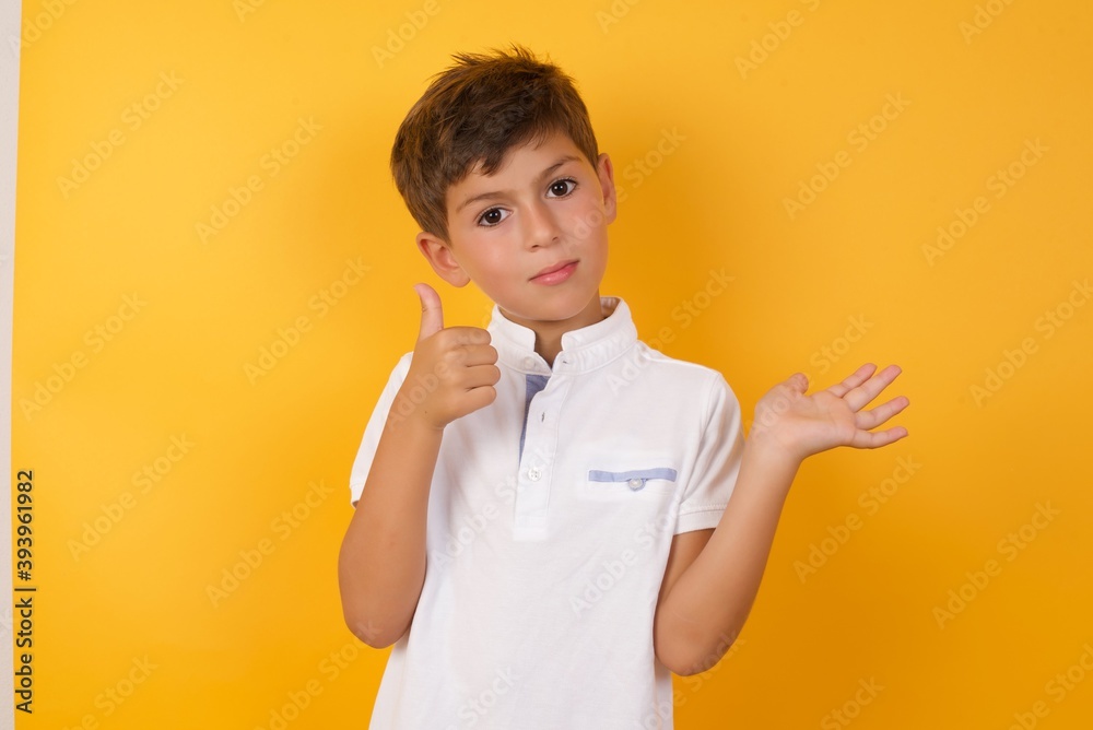 Happy cheerful little cute Caucasian boy kid wearing white t-shirt against yellow wall showing thumb up and pointing with the other hand. I recommend this. Happy cheerful little cute Caucasian boy kid wearing white t-shirt against yellow wall showing thumb up and pointing with the other hand. I recommend this.