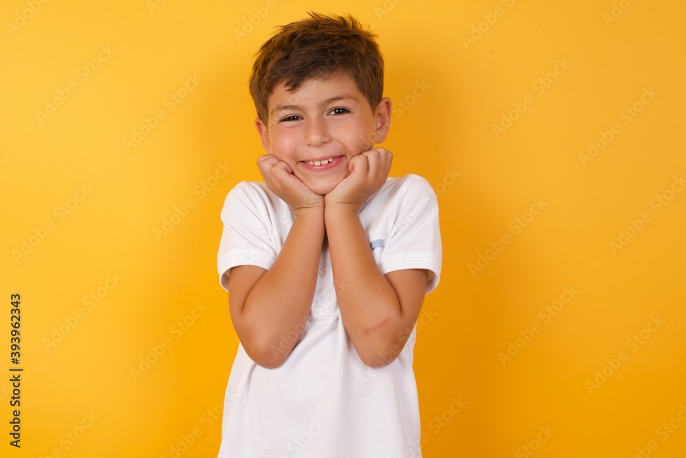 Portrait of little cute boy kid wearing white t-shirt against yellow wall being overwhelmed, expressing excitement and happiness with closed eyes and hands near face. Portrait of little cute boy kid wearing white t-shirt against yellow wall being overwhelmed, expressing excitement and happiness with closed eyes and hands near face.
