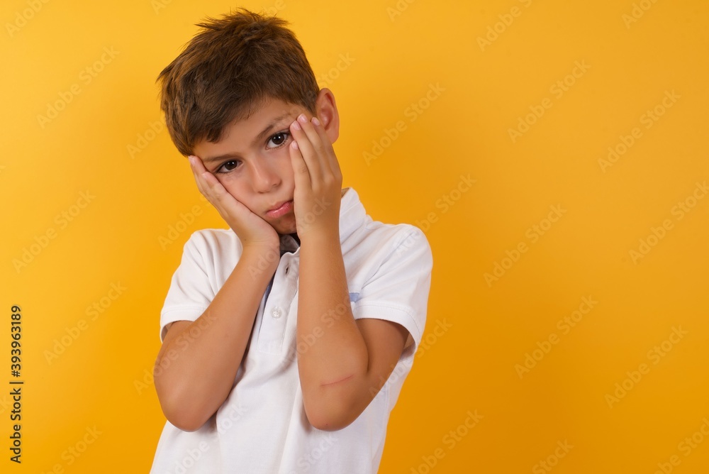 little cute boy kid wearing white t-shirt against yellow wall holding head in hands with unhappy expression watching sad movie about animals and trying not to cry. little cute boy kid wearing white t-shirt against yellow wall holding head in hands with unhappy expression watching sad movie about animals and trying not to cry.