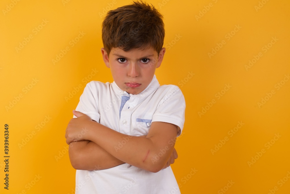 Picture of angry little cute boy kid wearing white t-shirt against yellow wall crossing arms. Looking at camera with disappointed expression. Picture of angry little cute boy kid wearing white t-shirt against yellow wall crossing arms. Looking at camera with disappointed expression.